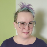 Head and shoulders photo of Andrea Steinwand, a non-binary caucasian person who is wearing an indigo coloured top. They have short and somewhat spiky two-toned hair, large frame glasses, a small silver labret piercing, and white stud earrings.
